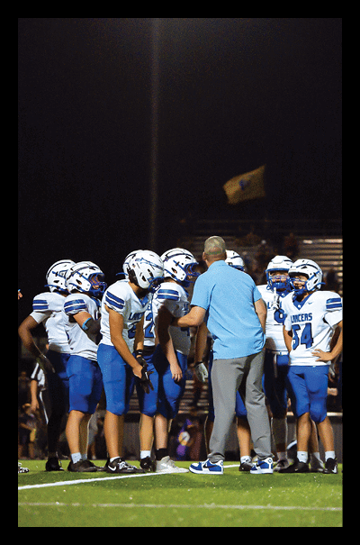 A group of people wearing blue and white football uniforms and helmets are gathered on a field at night with one person in a light blue shirt appearing to give instructions. Description generated by AI