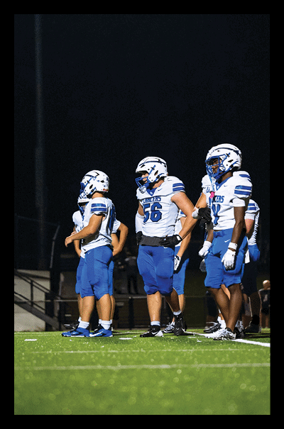 Several people wearing blue and white football uniforms and helmets stand together on a field at night. Description generated by AI