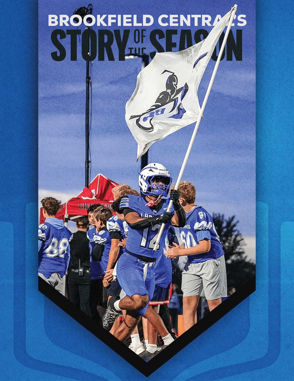 A person in a blue football uniform is running on a field holding a large white flag with a logo while other people in similar uniforms stand in the background under a twilight sky. Description generated by AI