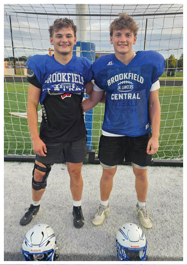 Two people wearing blue football shoulder pads and black shorts stand side by side on a sports field with a goal net behind them, one wearing a knee brace. Description generated by AI