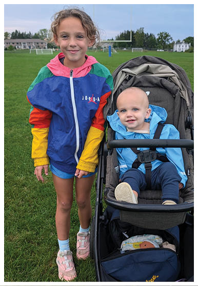 A child wearing a colorful jacket is standing next to a newborn in a stroller on a grassy field with houses and trees in the background. Description generated by AI