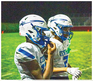 Two people wearing white and blue football helmets and jerseys are standing side by side, with one person adjusting their chin strap. Description generated by AI