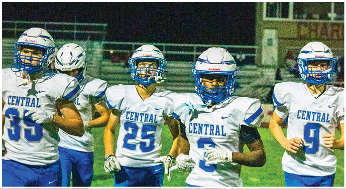 Four people wearing white and blue football uniforms with “CENTRAL" on the jerseys are walking together on a grassy field at night. Description generated by AI