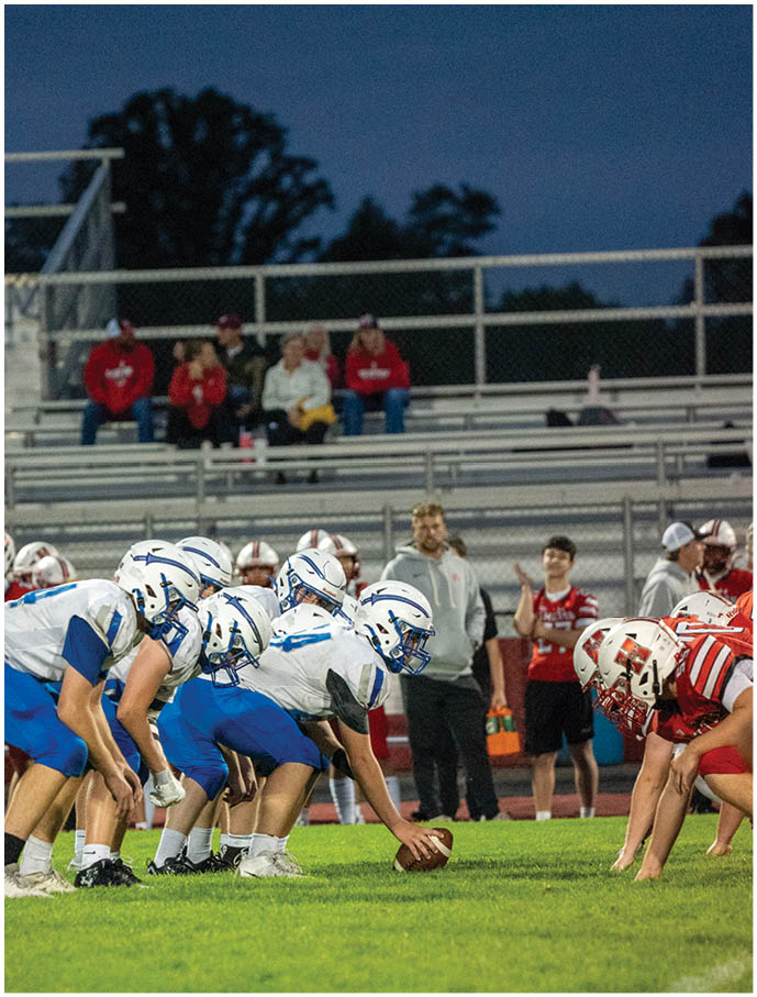 Two opposing American football teams in blue and white and red and white uniforms are lined up facing each other on a grassy field with spectators in the background during an evening game. Description generated by AI