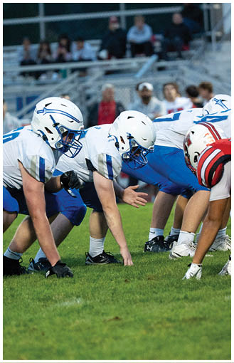Two people in white helmets and blue and white football uniforms are crouched in a ready position on a grassy field during a game. Description generated by AI