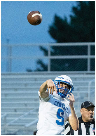 A person wearing a blue and white football uniform is throwing a football while a referee and several people in sports attire stand in the background near empty bleachers. Description generated by AI