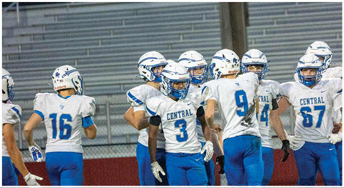 A group of people wearing white and blue football uniforms with helmets are gathered on a field near empty bleachers. Description generated by AI