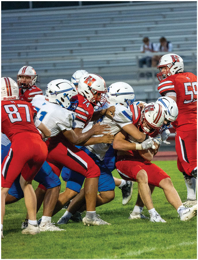A group of people wearing football uniforms are engaged in a tackle during a game on a grassy field with empty bleachers in the background. Description generated by AI