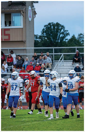 Several people in football uniforms stand on a grassy field with spectators seated in bleachers behind them. Description generated by AI