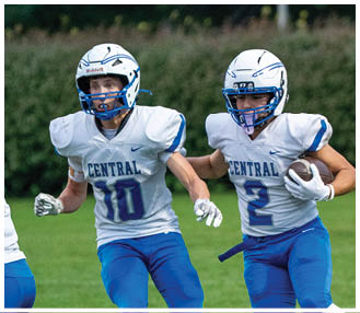 Two people in white and blue football uniforms are running on a grassy field with one holding a football while a referee and another player in red are visible in the background. Description generated by AI