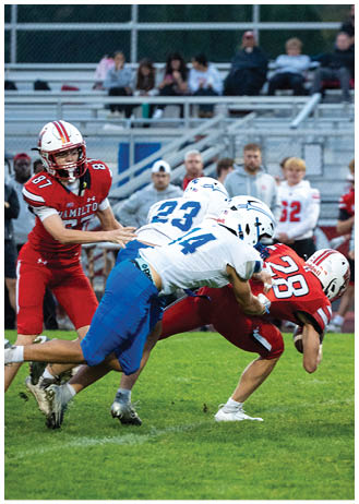 Two football players in red uniforms are being tackled by a player in a white and blue uniform on a grassy field with spectators in the background. Description generated by AI