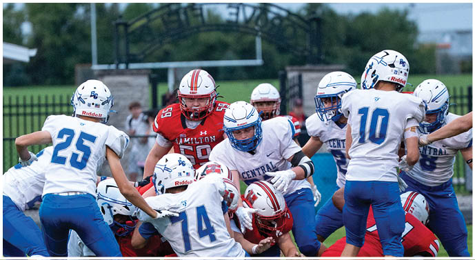 A group of people wearing football helmets and uniforms are engaged in a play on a grassy field with a fence and trees in the background. Description generated by AI