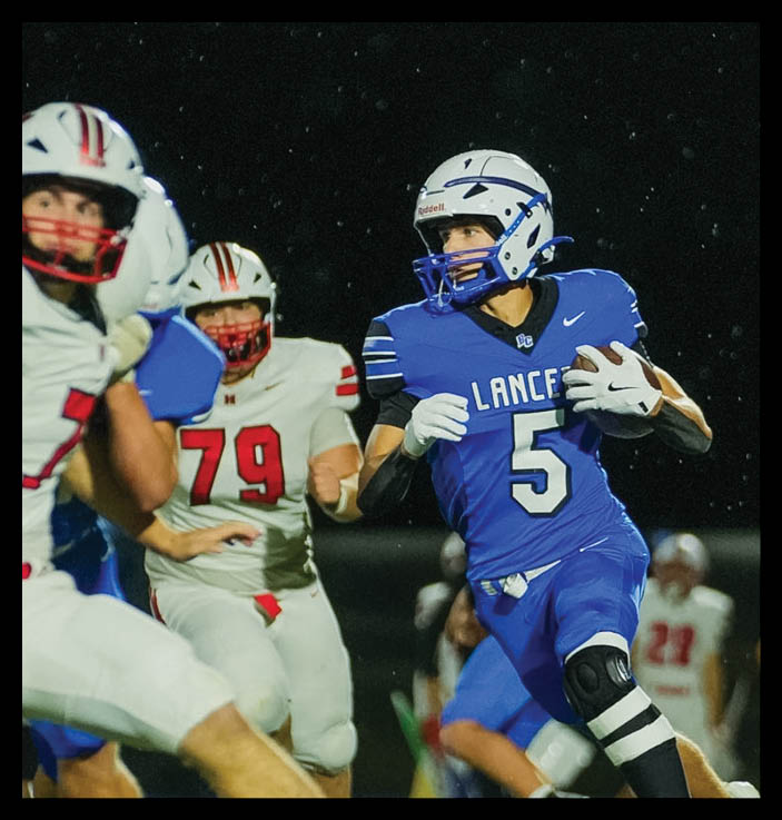 A person in a blue football uniform with the number 5 is running with the ball while people in white uniforms with red accents are attempting to catch them on a nighttime field. Description generated by AI