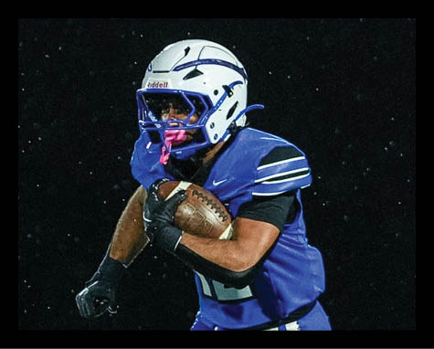 A person wearing a blue football uniform is running while holding a football tightly against their chest. Description generated by AI