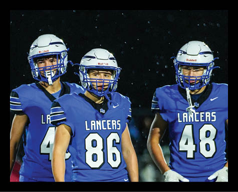 Three people wearing blue sports uniforms and white helmets stand closely together against a dark background with small light spots. Description generated by AI
