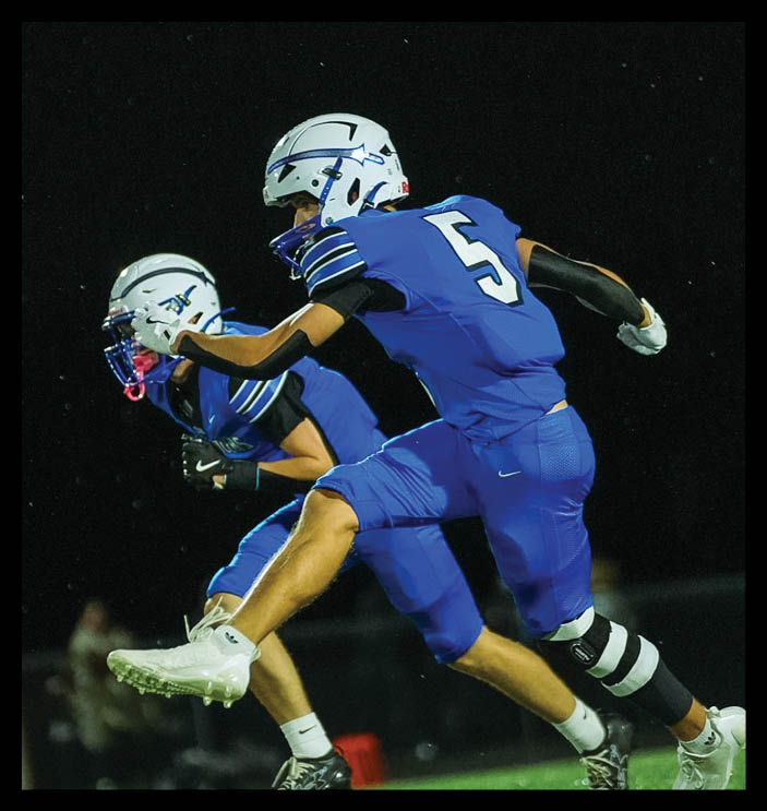 Two people wearing blue sports uniforms and helmets are engaged in a nighttime football game against a dark sky background. Description generated by AI