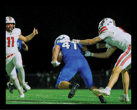 Two people in football uniforms are engaged in a play on a dark field, with one person in a blue jersey holding the ball and another in a white jersey reaching toward them. Description generated by AI