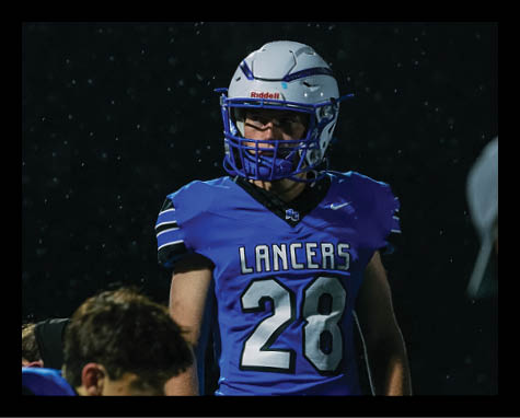 A person wearing a white football helmet with a blue faceguard is visible against a dark background with raindrops falling. Description generated by AI