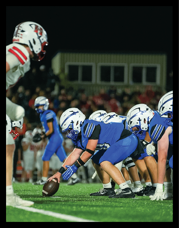 People in blue and white football uniforms are lined up on a field preparing for a play against opponents in white and red uniforms during a night game. Description generated by AI