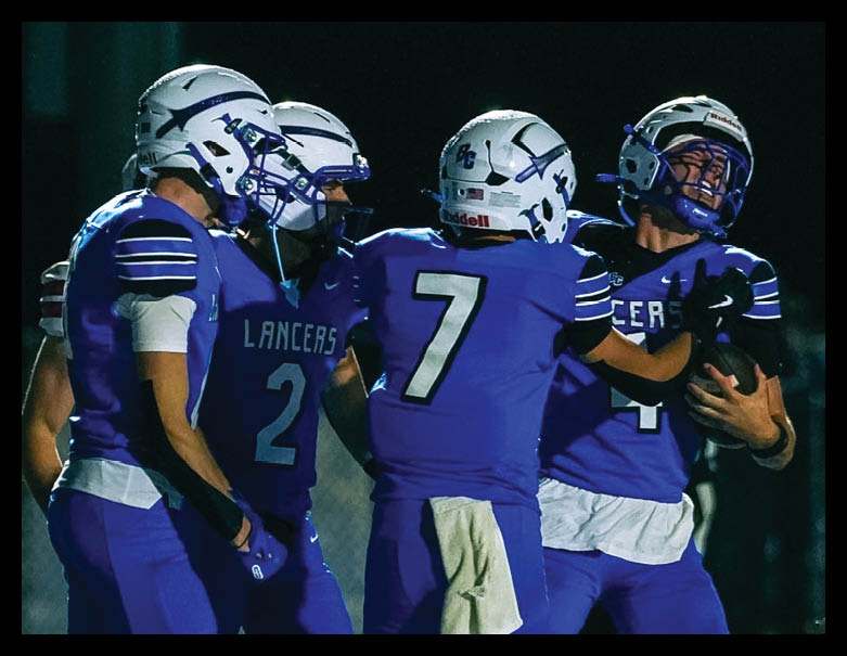 Four people wearing blue football uniforms and helmets are gathered closely together outdoors at night. Description generated by AI