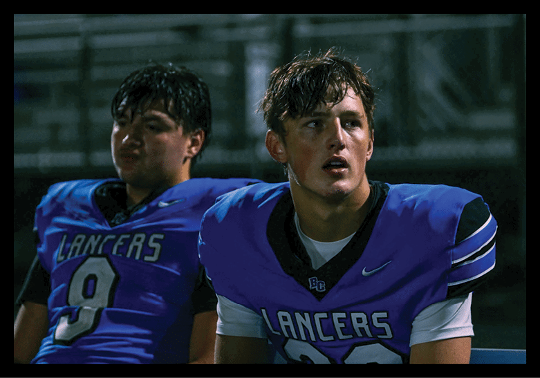 Two people wearing blue Lancers football uniforms sit on a bench with a dark background, one looking forward with a focused expression and the other looking to the side. Description generated by AI