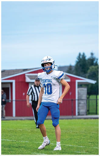 A person wearing a white and blue football uniform with the number 10 stands on a grassy field with a red building and a referee in the background. Description generated by AI