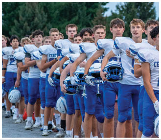 A line of people wearing blue and white football uniforms stand outdoors holding helmets in their arms. Description generated by AI