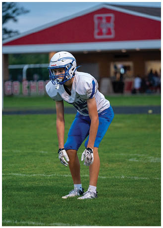 A person wearing a white football helmet, white jersey, and blue pants is standing on a grassy field in a ready position during a game. Description generated by AI