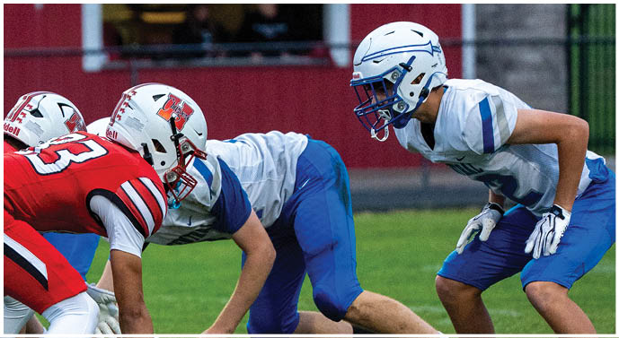Two opposing American football teams in red and white uniforms and blue and white uniforms are lined up facing each other on a grassy field preparing for a play. Description generated by AI