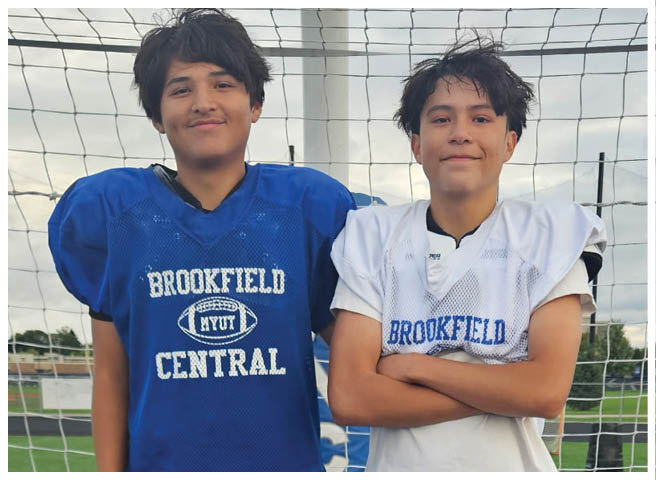 Two people wearing football jerseys stand side by side in front of a goal net outdoors with a cloudy sky in the background. Description generated by AI
