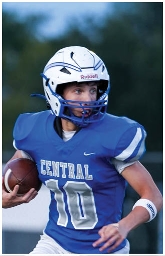 A person wearing a blue football jersey with the number 10 and a white helmet is holding a football and looking to the side during a game. Description generated by AI