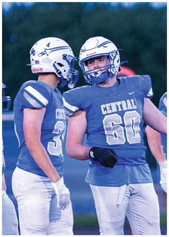 People wearing football uniforms and helmets are standing on a field, with one person in a blue jersey numbered 60 gesturing with their arm. Description generated by AI