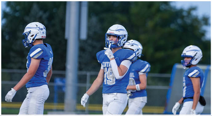 Four people wearing blue and white football uniforms and helmets stand on a field with trees and a pole in the background. Description generated by AI
