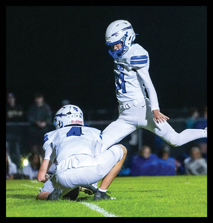 Two people in white football uniforms are on a grassy field at night with one person holding the ball on the ground and the other kicking it. Description generated by AI
