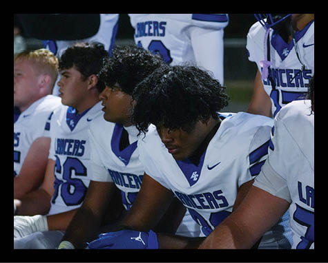 Several people in football uniforms sit closely together on a bench, appearing focused or contemplative. Description generated by AI
