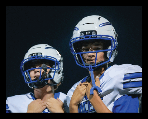 Two people wearing white and blue football helmets and uniforms stand close together against a dark background. Description generated by AI