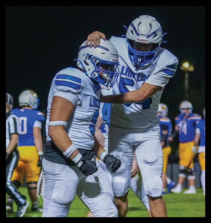 Two football players in white uniforms with blue accents celebrate on a field at night while other players and a referee stand in the background. Description generated by AI