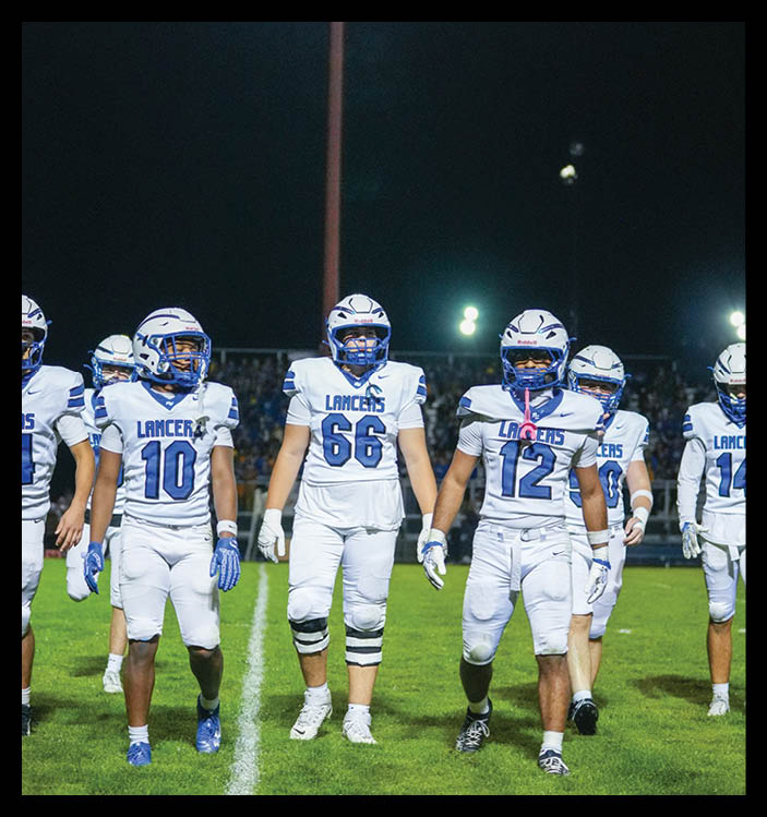 A group of football players in white uniforms with blue accents walk together on a grassy field at night under bright stadium lights. Description generated by AI
