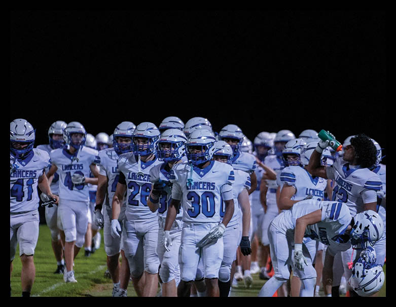 A group of football players in white and blue uniforms are walking together on a field at night, with one player drinking from a green water bottle. Description generated by AI