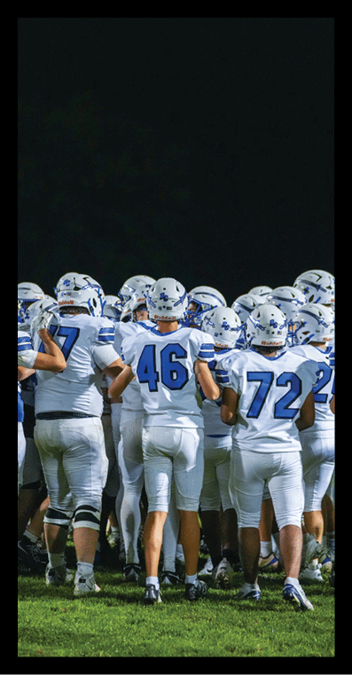 A group of people wearing white football uniforms with blue numbers are gathered closely together on a grassy field at night. Description generated by AI