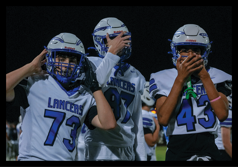 Three people wearing white football uniforms with blue helmets and the word LANCERS on their jerseys stand together on a field at night, with two of them holding their helmets and one covering their mouth. Description generated by AI
