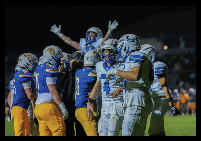Players from two opposing football teams in blue and white uniforms are gathered on the field at night with one player raising their arms in celebration. Description generated by AI