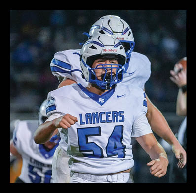 A group of football players in white and blue uniforms celebrate on a field with one player in the foreground pumping a fist. Description generated by AI