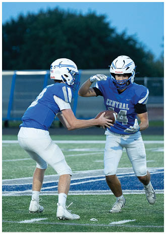 Two people in blue football uniforms and helmets are engaged in a play with one handing the football to the other on a field with a blurred background of trees and a fence. Description generated by AI