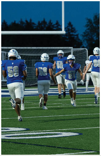 People wearing blue and white football uniforms are running on a green field toward a goalpost during a game or practice at dusk. Description generated by AI