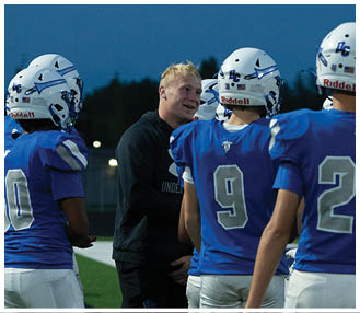 Several people wearing blue sports jerseys and white pants are standing on a green field, likely during a football game or practice. Description generated by AI