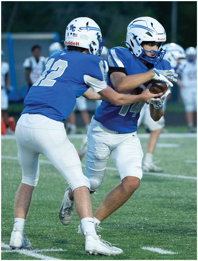 Two people wearing blue and white football uniforms are on a field with one handing off the ball to the other while other players stand in the background. Description generated by AI