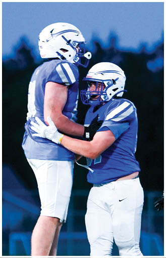 Two people wearing football helmets and uniforms are embracing on a field with a dark background. Description generated by AI