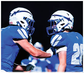 Two people wearing blue football uniforms and white helmets face each other against a dark background. Description generated by AI