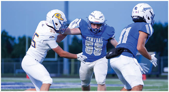 Two people in football uniforms are engaged in a play on a field, with one wearing a blue jersey labeled “Central" and the other in a white jersey with a helmet featuring a yellow and blue logo. Description generated by AI